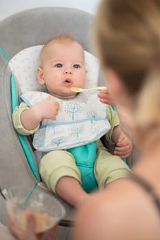Mother spoon feeding her baby boy child in baby chair with fruit puree. Baby solid food introduction concept