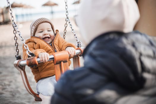 Mother pushing her cheerful infant baby boy child on a swing on sandy beach playground outdoors on nice sunny cold winter day in Malaga, Spain