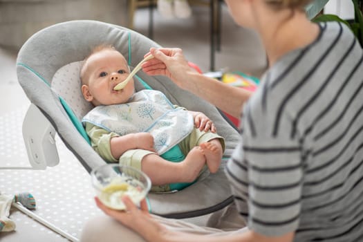 Mother spoon feeding her baby boy child in baby chair with fruit puree. Baby solid food introduction concept