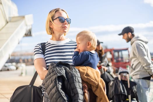 Motherat travelling with his infant baby boy child. Mom holding travel bag and her infant baby boy child while queuing for bus in front of airport terminal station
