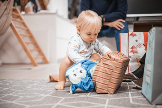 Cute little blond Caucasian toddler unpacking Christmas or Birthday present. Adorable small infant baby boy opening gift.