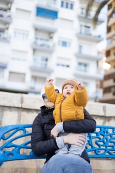 Young mother with her cute infant baby boy child on bench in city park