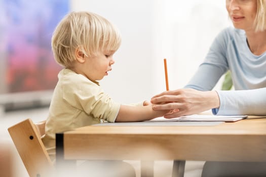 Caring young Caucasian mother and small son drawing painting in notebook at home together. Loving mom or nanny having fun learning and playing with her little 1,5 year old infant baby boy child