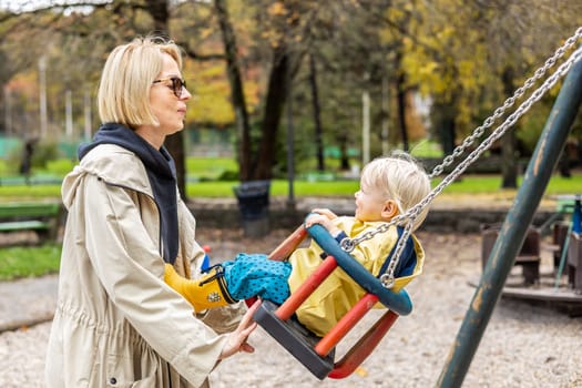 Mother pushing her infant baby boy child wearing yellow rain boots and cape on swing on playground outdoors on cold rainy overcast autumn day in Ljubljana, Slovenia.