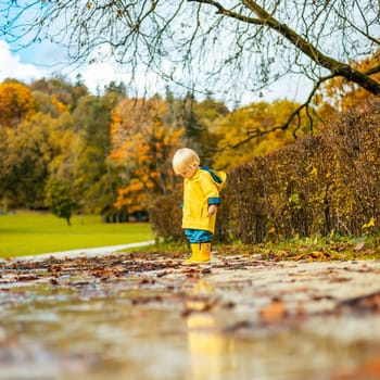Sun always shines after the rain. Small bond infant boy wearing yellow rubber boots and yellow waterproof raincoat walking in puddles in city park on sunny rainy day