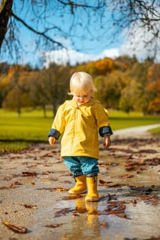 Sun always shines after the rain. Small bond infant boy wearing yellow rubber boots and yellow waterproof raincoat walking in puddles in city park on sunny rainy day