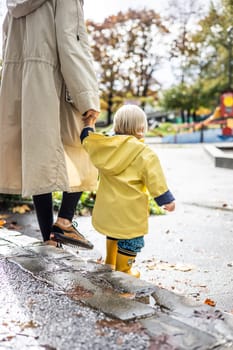 Small bond infant boy wearing yellow rubber boots and yellow waterproof raincoat walking in puddles on a overcast rainy day holding her mother's hand. Mom with small child in rain outdoors