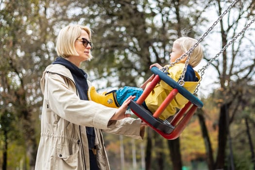 Mother pushing her infant baby boy child wearing yellow rain boots and cape on swing on playground outdoors on cold rainy overcast autumn day in Ljubljana, Slovenia.