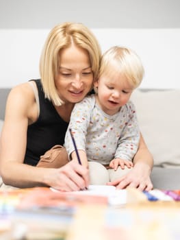 Caring young Caucasian mother and small son drawing painting in notebook at home together. Loving mom or nanny having fun learning and playing with her little 1,5 year old infant baby boy child