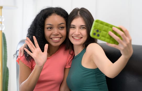Two smiling young women taking a selfie with mobile phone at home. Caucasian girl in green t-shrit holding smartphone and African female raise a hand waving while sitting on couch.