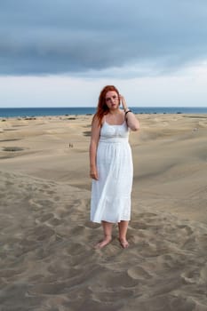 A young girl stands in a white dress in the sand dunes under a cloudy sky
