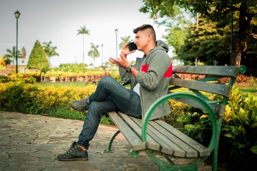 Man sitting on a bench calling on the phone, young man sitting on a bench with cell phone, Man calling on the phone sitting on a bench in the park