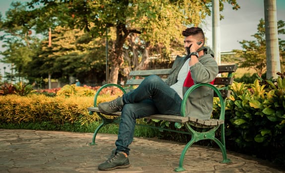 Man sitting on a bench calling on the phone, young man sitting on a bench with cell phone, close up of a young man calling on the phone sitting on a bench