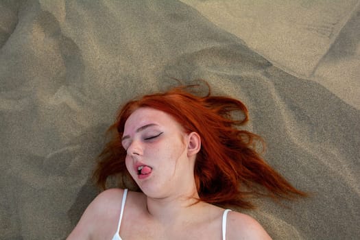 A young girl lies in the sand dunes in a white dress, with a funny facial expression, Tongue outside