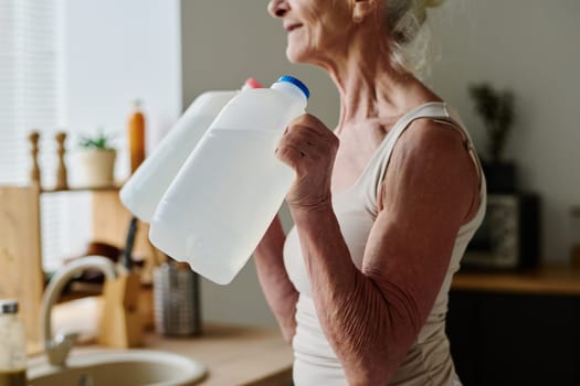 Cropped shot of senior woman in activewear doing physical exercise with two plastic canisters with water while standing in front of camera