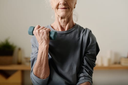 Cropped shot of senior active woman in grey cotton pullover sitting in front of camera at home and practicing exercise with dumbbells