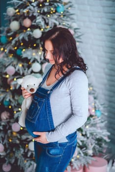 A pregnant brunette girl in a denim blue jumpsuit with a teddy bear stands near the Christmas tree in the studio