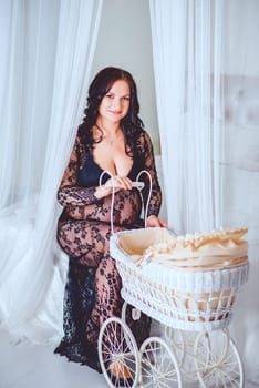 A pregnant brunette girl in a black lace dress poses with a stroller sitting on a bed with white curtains in the studio