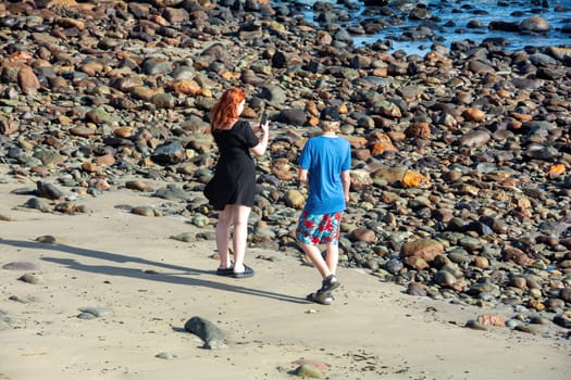 Young woman wearing a short black dress stands on a stony beach and takes photos with her cell phone, a boy stands next to her