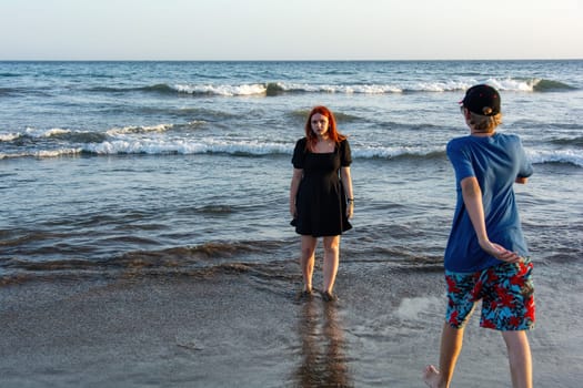 A girl in a black summer dress stands in the waves by the sea, a boy in shorts and a cap runs to her