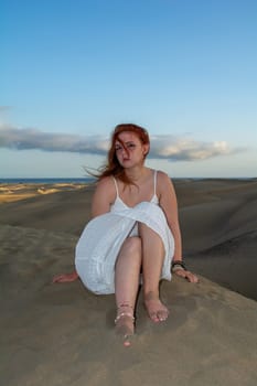 A young pretty red-haired girl in a white dress sits in the sand dunes in the late afternoon and enjoys the last rays of sunshine
