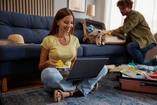 Wife booking ticket paying for hotel using laptop and bank credit card while husband preparing luggage bag for trip adventure. Planning vacation travel concept