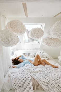Dreamy relaxed woman in bed lying and looking up on fluffy decoration in shape of clouds on ceiling. Attractive romantic female dreamer rest in cozy room on attic floor