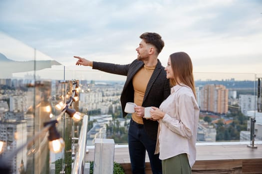 Business colleagues chitchatting during coffee break at office roof terrace. Young man project manager pointing to city building discussing real estate