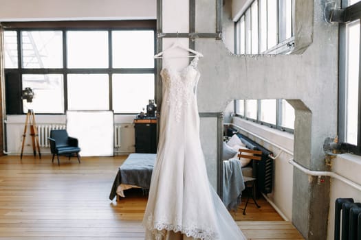 A white long wedding dress is hanging on a hanger in a larger studio