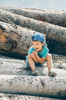 A little boy in a blue baseball cap and short shorts sits on a log during the day and laughs merrily