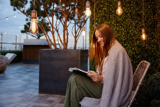 Young woman wrapped in warm blanket reading book enjoying relaxation in cozy place of home terrace or outdoor cafe. Romantic dream mood and imagination