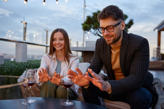 Relaxed young romantic couple warming hands at decorative fireplace while having date on rooftop. Boyfriend and girlfriend drinking wine enjoying pastime together