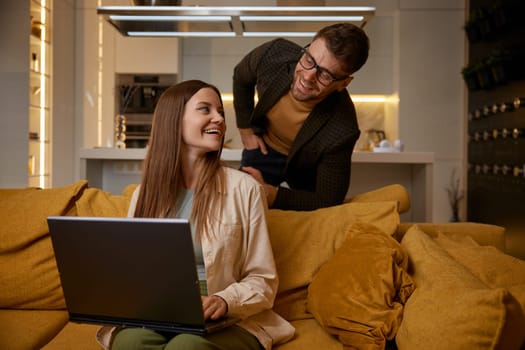 Happy stylish married couple resting at home. Young man and woman having fun in social media, watching funny video on laptop computer sitting on sofa in living-room. Weekend leisure