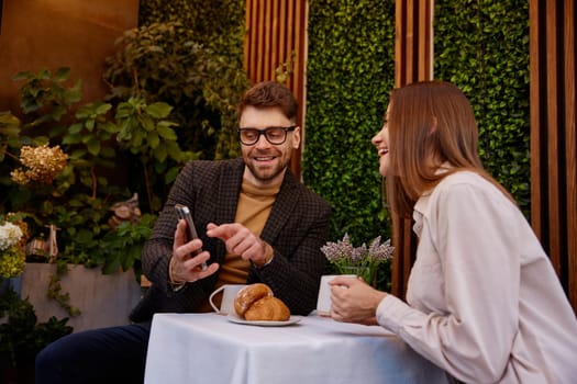 Young man and woman talking using mobile phone having business meeting in cafe. Carefree businesspeople viewing social media networks enjoying relaxing moment during work break