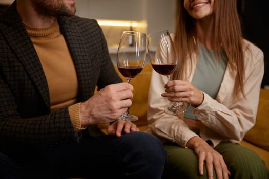 Romantic evening at home with closeup view on clinking red wine glasses in hands of male and female. Happy young couple enjoying nice idyllic atmosphere at home, celebrating start of weekends