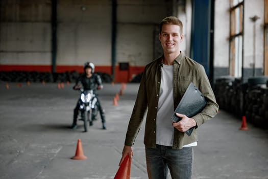 Portrait of happy smiling instructor of driving school at training motordrome with motorcyclist student riding motorbike on background