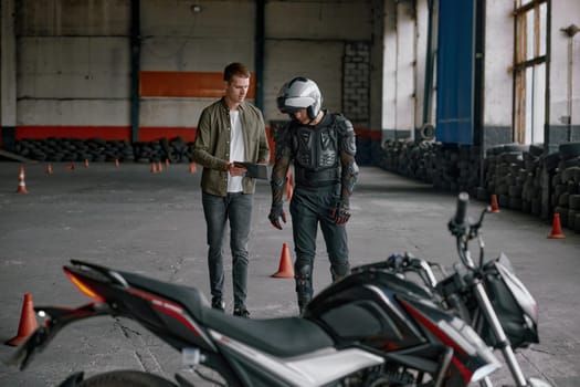 Student motorcyclist and instructor discussing rout of future exam race. Two men standing together looking at checklist at indoor motordrome of driver training center