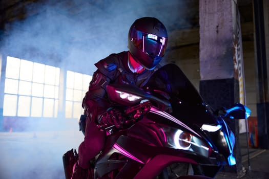 Portrait of motor biker wearing protective suit and helmet headset driving sport motorcycle in neon light and smoke. Indoor motordrome for speed riding practice