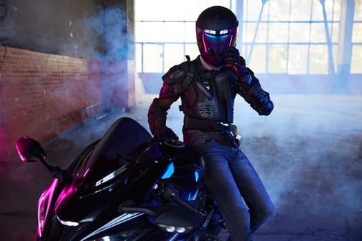 Portrait of man extreme rider sitting at motorcycle taking rest after training at driving school motordrome. Atmospheric photo in neon light illumination and smoke