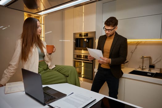 Young family discussing income and evaluating expenditure at home kitchen. Husband reading bill tax, woman with cup of coffee listening attentively