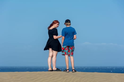 Two teenagers stand on a sand dune, with sea and blue sky