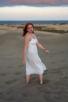 A young girl stands in a white dress in the sand dunes with sky and clouds in the late afternoon