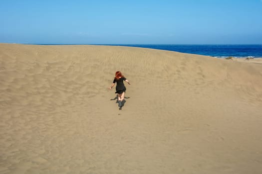 Young woman running up a sand dune towards the sea, with blue sky