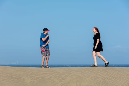 Two teenagers stand on a sand dune, with sea and blue sky