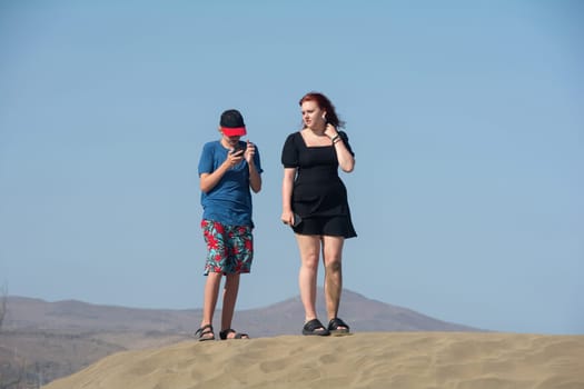 Two teenagers are standing on a sand dune with their cell phones, the boy is looking at his smartphone