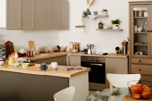Part of modern spacious kitchen with row of cabinets hanging over counter with kitchenware and sink and electric stove with coffee pot