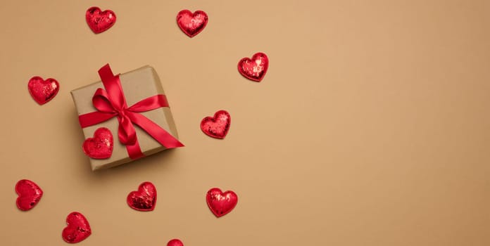 Box tied with a red ribbon and small hearts on a beige background, top view. Copy space