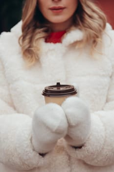 Woman in white wool mittens and fur coat holding take away coffee cup outdoors, soft focus on hands.