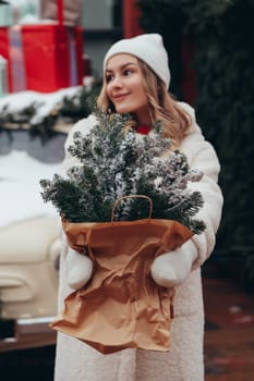 Woman in white hat and winter fur coat with pine branch in city, lights and decorations on background.