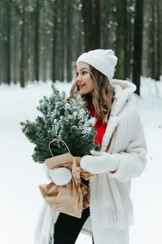 Young blonde woman wears white fur coat, knitted hat and mittens, posing with fir tree branches wrapped with craft paper in winter forest. Winter fashion